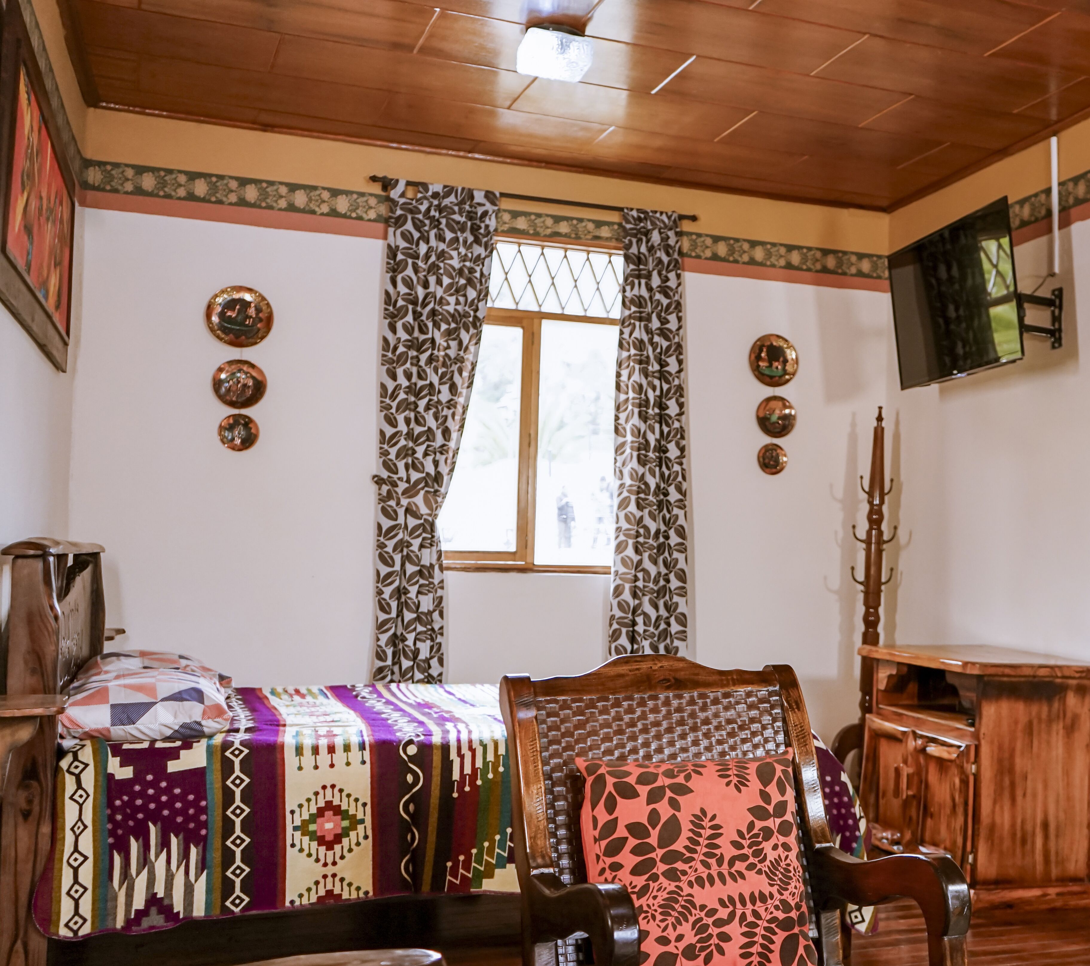A room with a wooden ceiling, a bed with patterned bedding, a chair, a wooden cabinet, and a window with curtains.