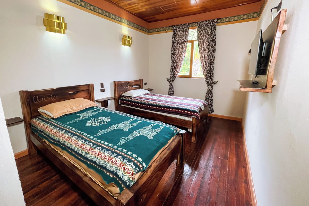 A bedroom with two wooden beds, a wooden ceiling, a small TV on a wall, and a window with patterned curtains.
