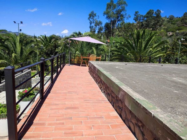 Un sendero de ladrillos conduce a un área de descanso con una mesa y sillas, rodeada de abundante vegetación y un cielo azul despejado.