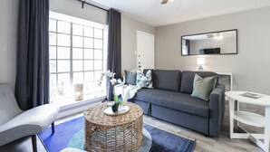 Een moderne woonkamer met een grijze bank, een ronde salontafel van riet en een groot raam met gordijnen.