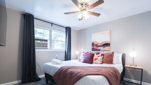 Een slaapkamer met een bed, een plafondventilator, een nachtkastje met een lamp, een raam met gordijnen en een schilderij aan de muur.