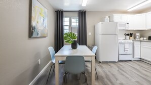 Een moderne keuken met witte kastjes, een witte koelkast, een magnetron en een fornuis. Er is een eettafel met twee stoelen en een potplant.
