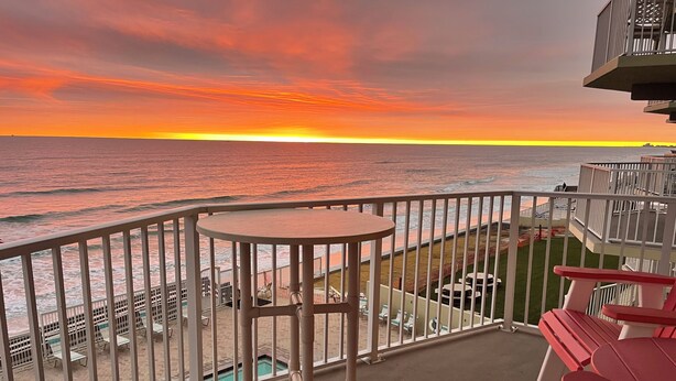 Un balcón con mesa y sillas, con vista a una piscina y una playa al atardecer.