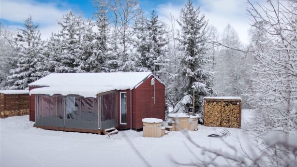A snow-covered outdoor area with a red building, possibly a cabin or lodge, surrounded by snow-laden trees. There are several wooden benches and a stack of firewood nearby. - Pod Liščím kopcem 2+2 by Interhome (Dolni Branna)