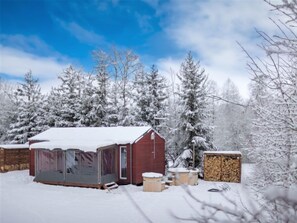 A snow-covered outdoor area with a red building, possibly a cabin or lodge, surrounded by snow-laden trees. There are several wooden benches and a stack of firewood nearby.