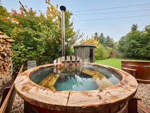 A hot tub with a wooden exterior, surrounded by a garden with trees and a wooden structure in the background.