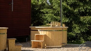 A wooden hot tub with a metal pole, surrounded by wooden benches and a wooden structure, set against a backdrop of trees and a red building. - Pod Liščím kopcem 2+2 by Interhome (Dolni Branna)