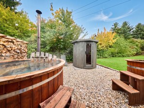 A hot tub with a wooden deck, a sauna, and a small wooden cabin in a garden setting.