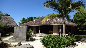 A traditional building with a tiled roof, a thatched-roof structure, and a garden with a palm tree and flowering plants. - ADAN RESORT -LANTERN- (Motobu)