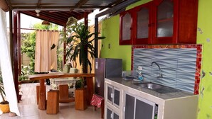 Een kleine keuken met een spoelbak, een koelkast en een fornuis. Er is een houten tafel met stoelen, en potplanten.