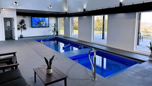 An indoor swimming pool with clear views of the outdoors, a large TV screen, and a seating area with a table and chairs.