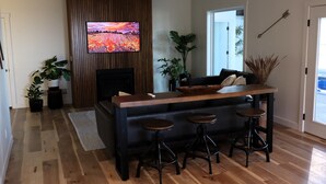 A modern living room with a wooden floor, a TV mounted on the wall, a sofa, a coffee table, and two bar stools.