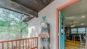A wooden deck with a view of trees, a bear sculpture labeled "APACHE PARADISE", and a teal door leading inside.