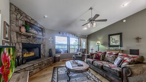 A living room with a stone fireplace, a ceiling fan, and a large window offering a view of the outdoors.