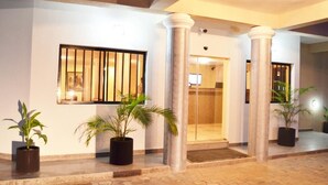 Entrada de un hotel con puerta de vidrio, plantas en macetas y suelo de baldosas.