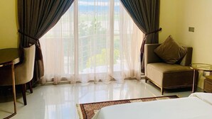Habitación de hotel con cama, una silla, un escritorio y vista a zonas verdes a través de cortinas translúcidas.