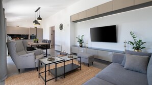 Uma sala de estar moderna com um sofá cinzento, cadeiras com padrão preto e branco, uma mesa de centro de vidro e uma TV de tela plana na parede.