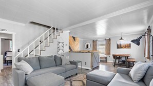 Un salon moderne avec un canapé d’angle gris, un mur en bois orné d’un motif en chevrons, un escalier et un coin repas comprenant une table et des chaises en bois.