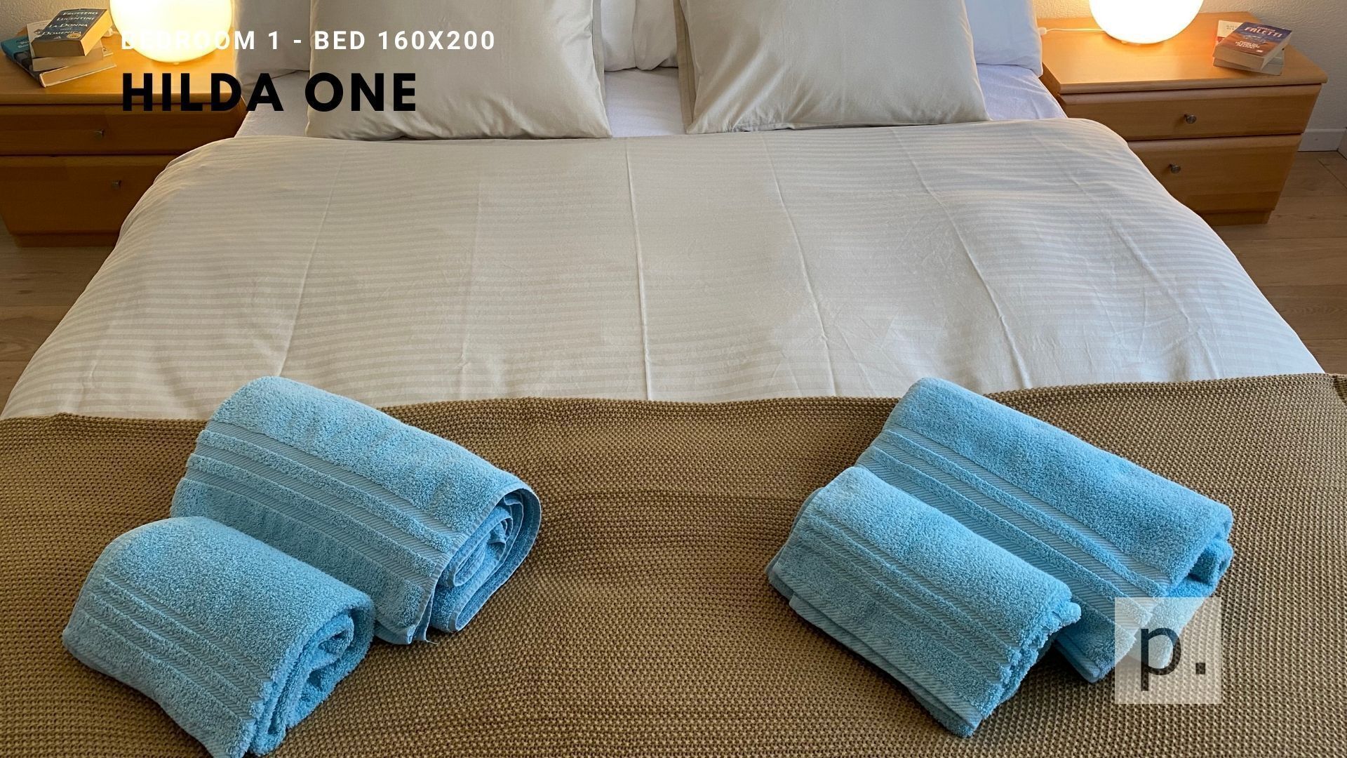 Une chambre d’hôtel avec un grand lit, deux oreillers, une table de chevet avec une lampe, et deux serviettes pliées posées sur un tapis marron.
