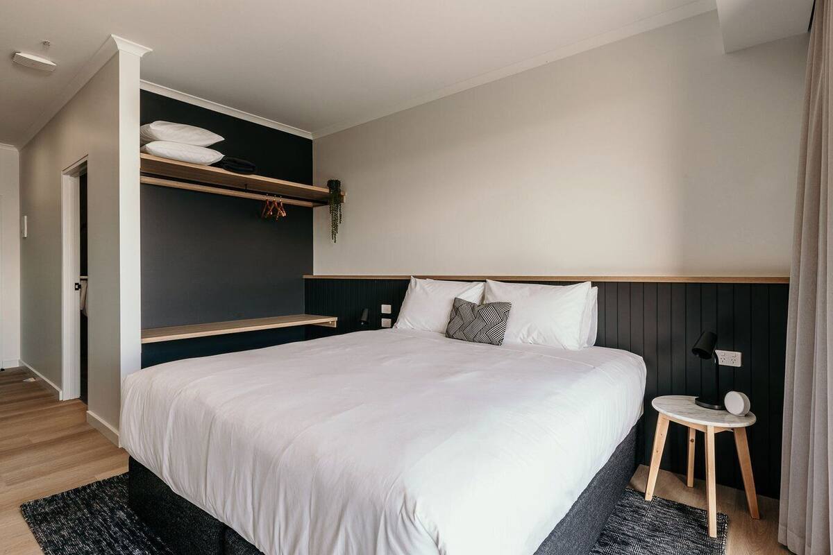 A modern hotel room with a large bed, a bedside table, a built-in shelf with pillows, and a small side table with a lamp.
