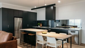 Une cuisine moderne avec des meubles de couleur sombre, des appareils en acier inoxydable et une table à manger en bois, assortie de chaises blanches.