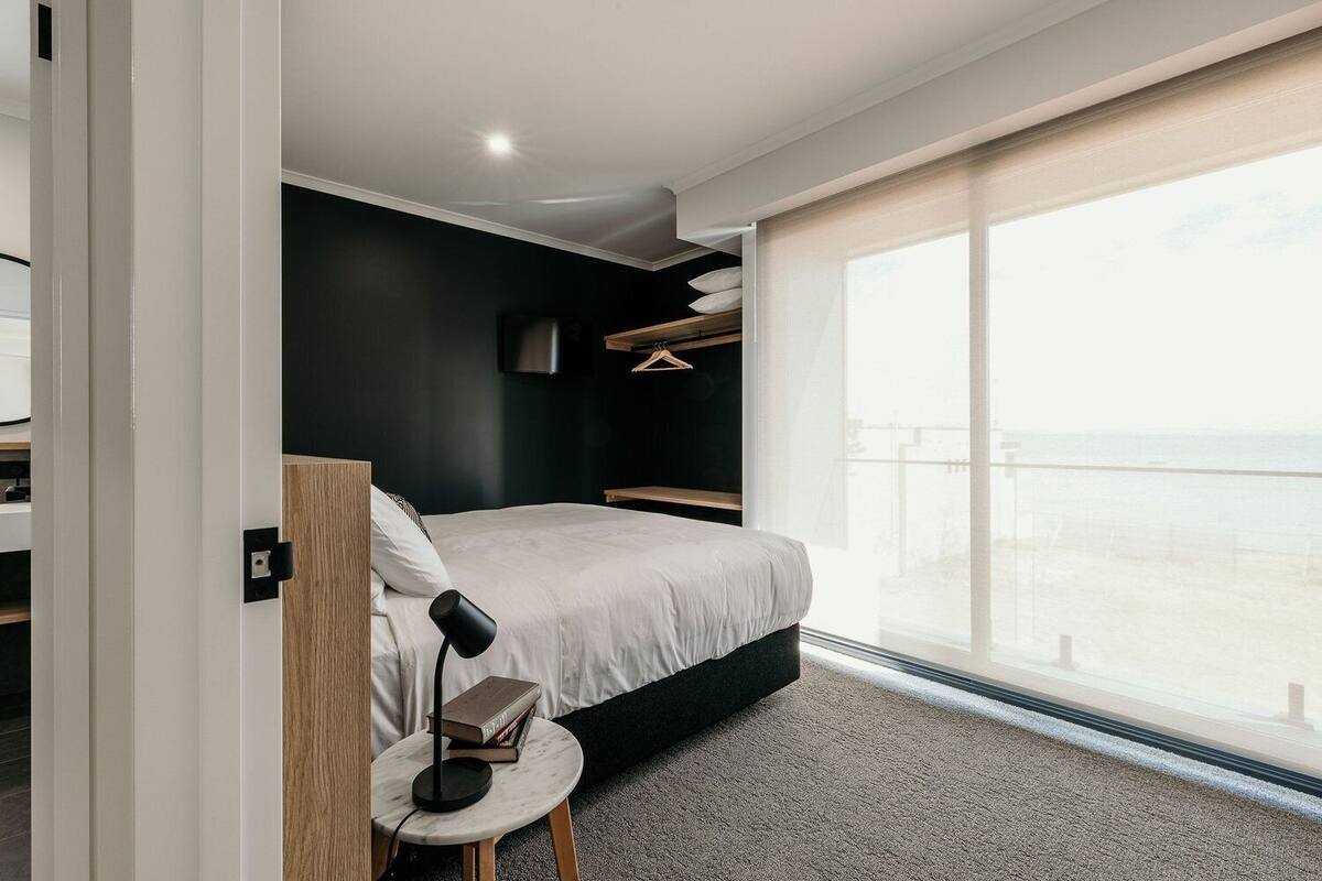 A modern bedroom with a large window, a bed with a white mattress, a bedside table with a lamp and books, and a TV mounted on the wall.