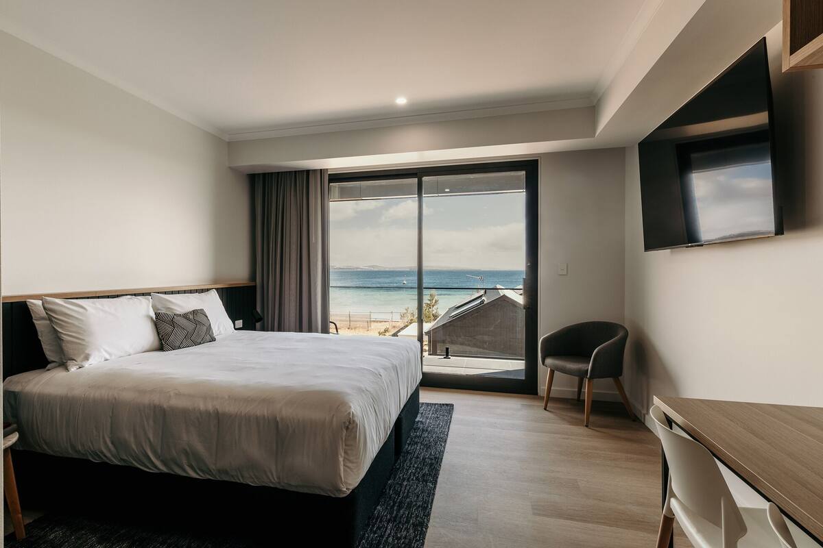 A modern hotel room with a large bed, a desk, and a view of the sea.