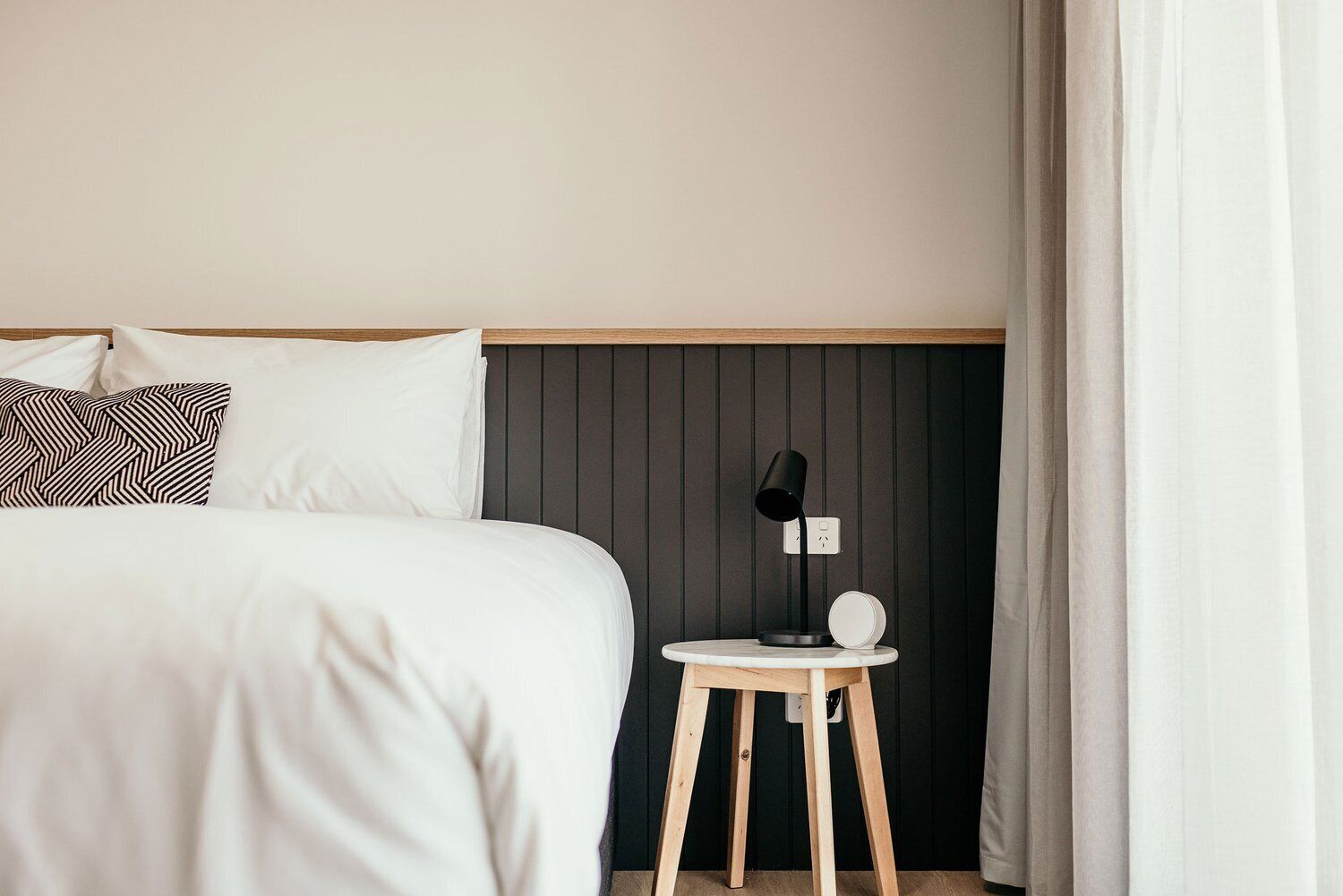 A neatly made bed with white linens and patterned pillows, a bedside table with a lamp, and a dark wooden panel above the bed.