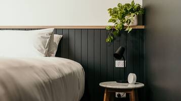 Ein ordentlich bezogenes Bett mit weißen Leinentüchern, ein kleiner runder Tisch mit einer Lampe und eine Topfpflanze auf einem Regal über dem Bett.