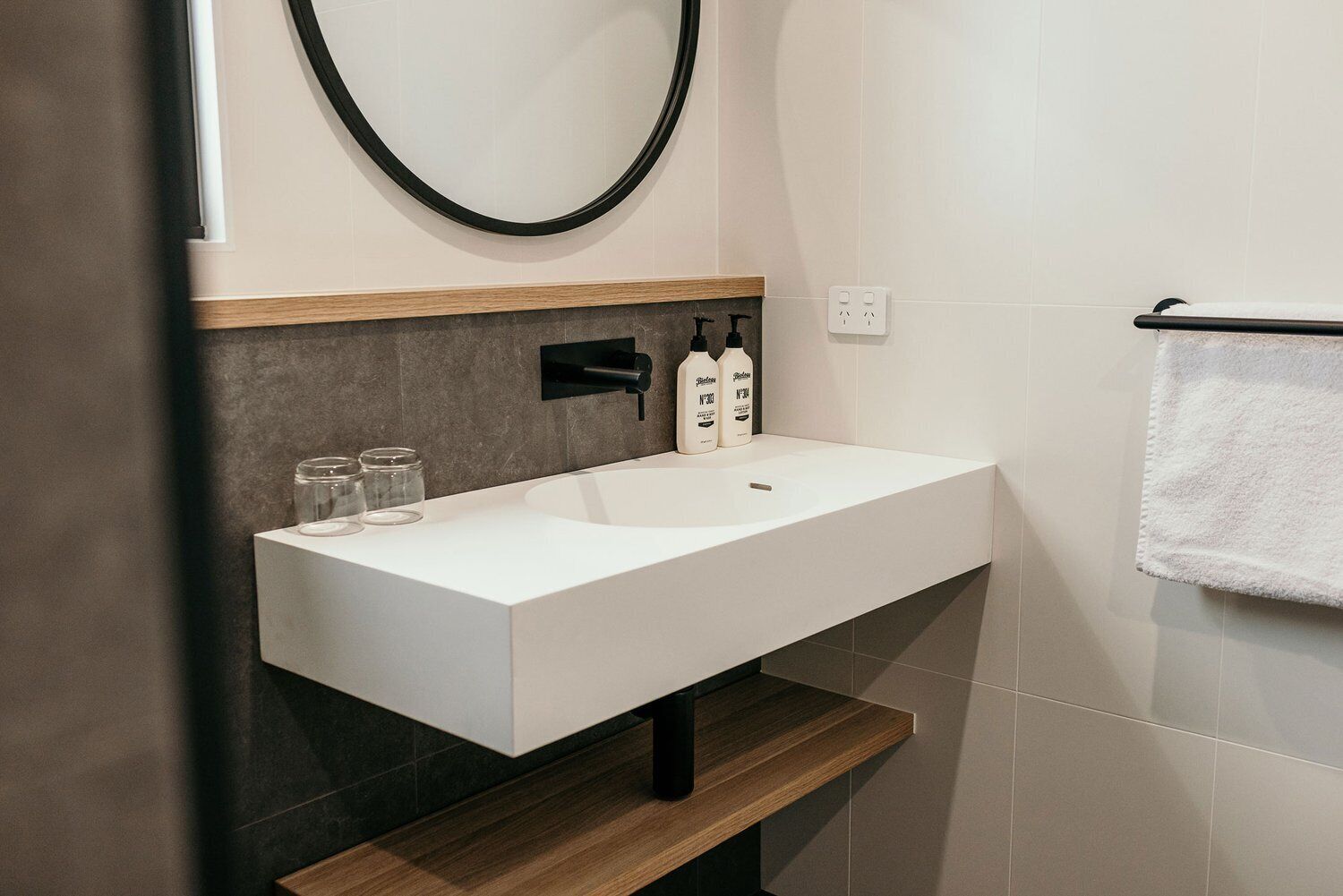 Un baño moderno con un lavamanos blanco, baldosas oscuras, un estante de madera y un perchero.