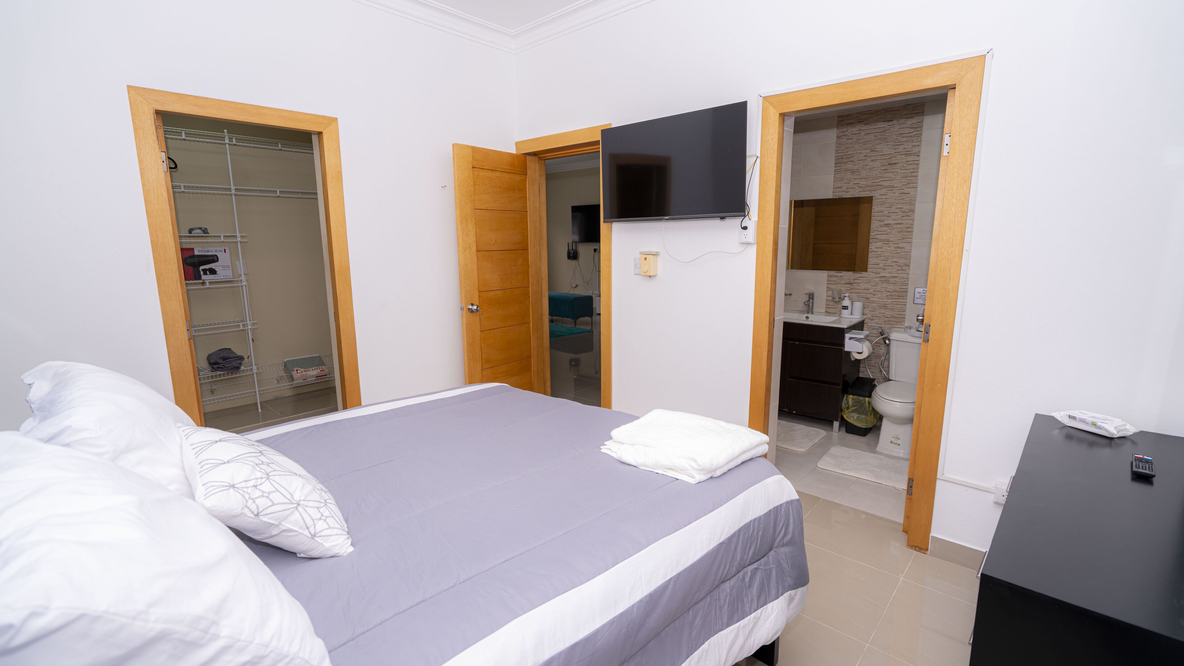 Un dormitorio con cama, televisor montado en la pared y baño visible a través de una puerta abierta.