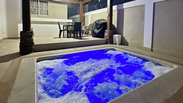 Ein Schwimmbecken mit blauer Beleuchtung, eine überdachte Terrasse mit Stühlen und eine Steinsäule.
