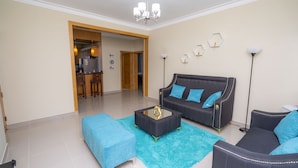 Ein Wohnzimmer mit einem dunkelgrauen Sofa, blauen Kissen, einem Glastisch und einem türkisgrünen Teppich. Durch einen offenen Durchgang ist eine Küche erkennbar.
