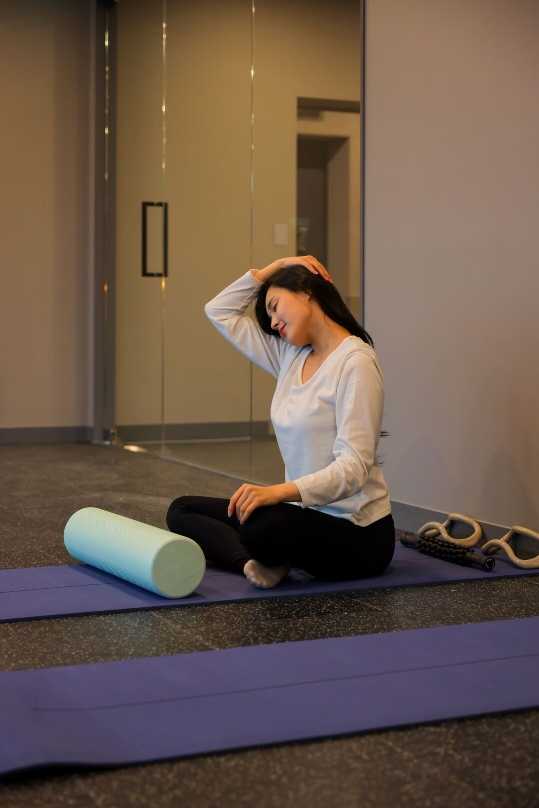 Eine Person sitzt auf einer violetten Yogamatte in einem Raum mit Glaswand und Tür im Hintergrund.