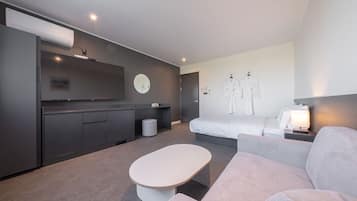 Quarto de hotel moderno com sofá cinzento, mesa de centro branca, cama com cabeceira branca, pequena mesa de cabeceira com abajur, televisão na parede e um armário cinzento.