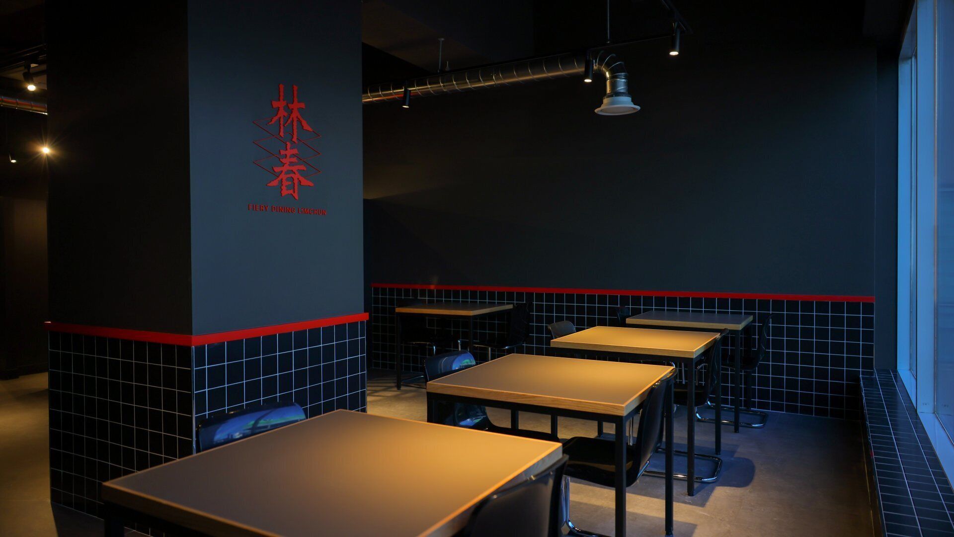 A restaurant interior with tables and chairs, a dark wall with red accents, and a logo on the wall.