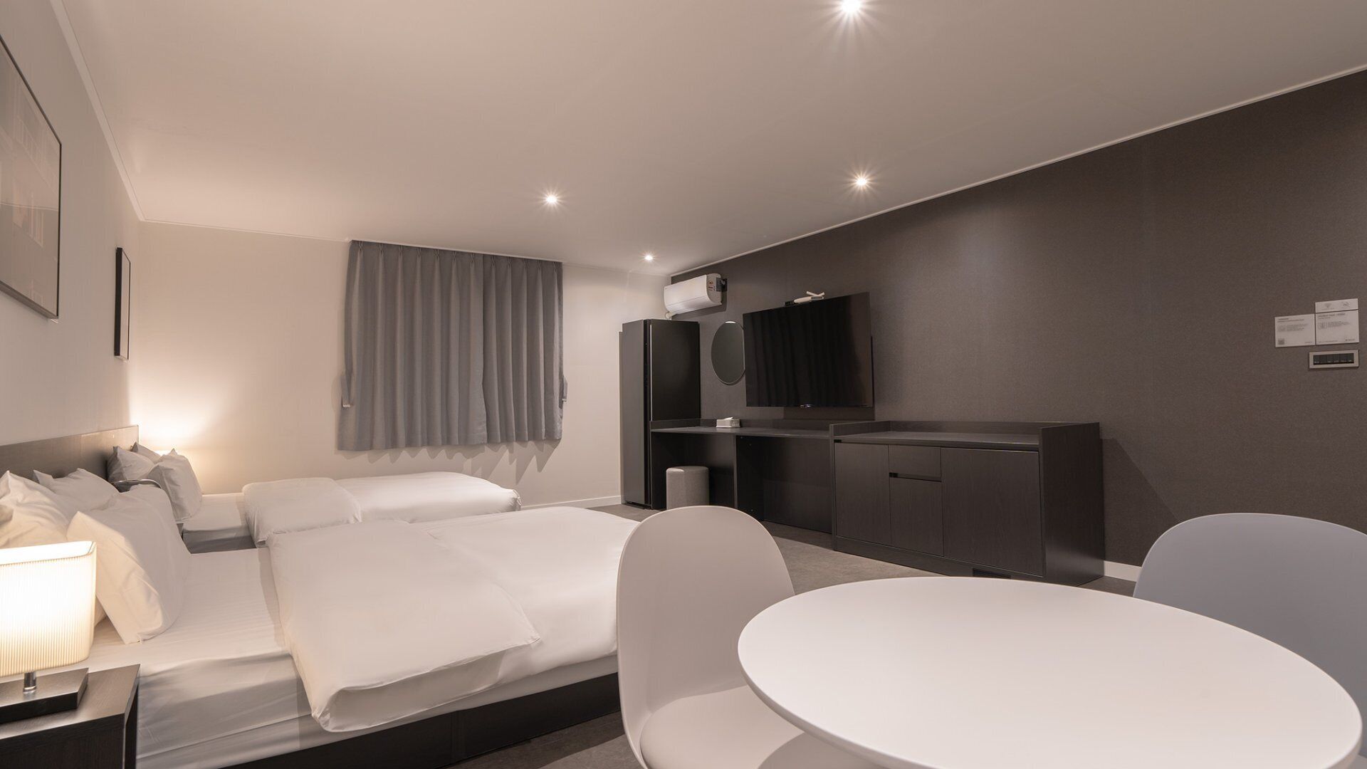 A modern hotel room with a bed, bedside table, lamp, and a round dining table with chairs.
