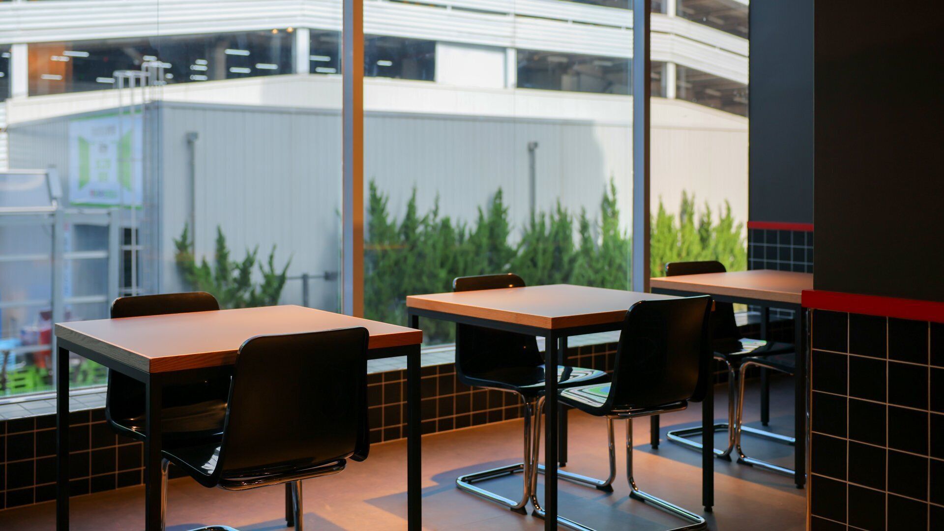 A modern dining area with tables and chairs, large windows, and a view of greenery outside.