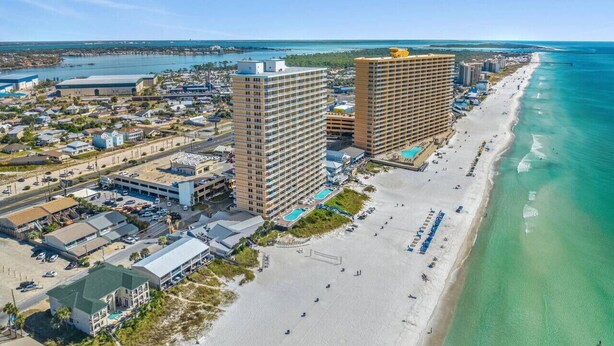 A coastal cityscape with a beachfront hotel featuring multiple floors, a parking lot, and a sandy beach. - Seychelles 703 | Beach Chair Service Included at this 7th Floor, Oceanfront Condo (Panama City Beach)
