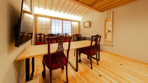 Een eethoek met een lange houten tafel, rode stoelen met kussens, een aan de muur bevestigde televisie en een groot raam met houten kozijnen.