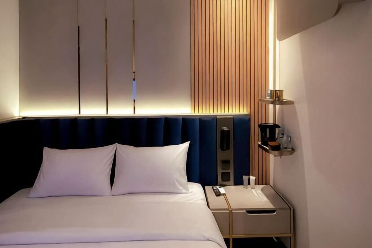 A modern hotel room with a bed, bedside table, and a wall-mounted shelf with a cup and a small decorative item.