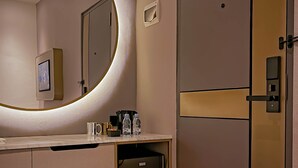 A modern bathroom with a vanity, a large mirror, a toilet, and a small stool. - Qube Hotel by 98hospitality (Jakarta)