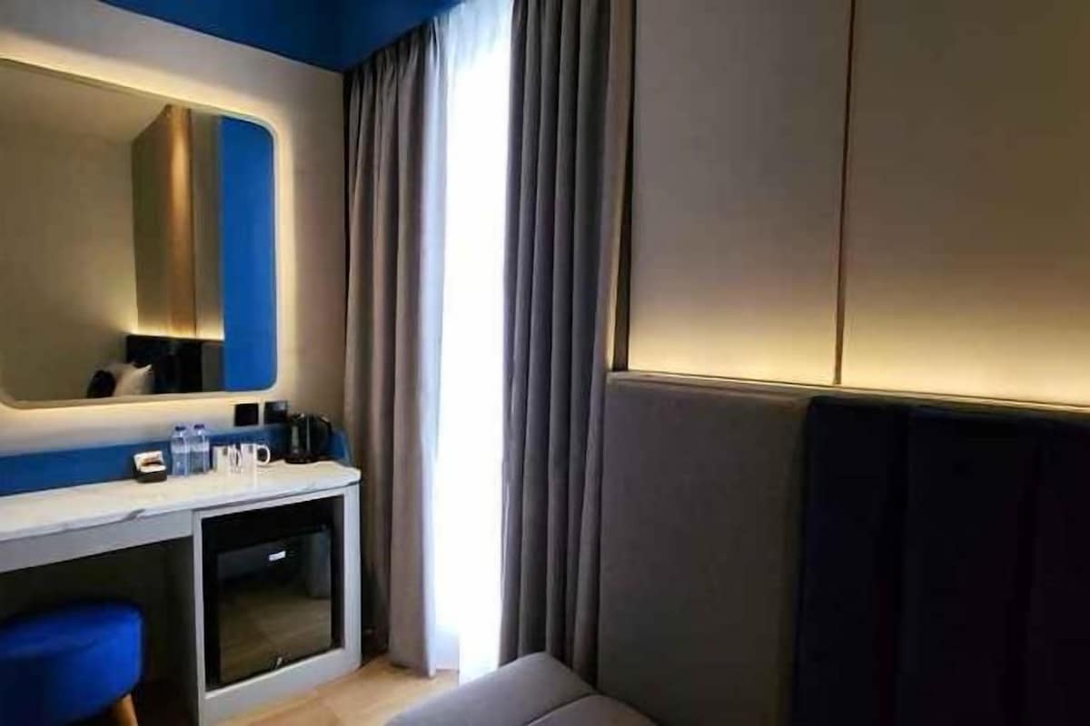 Una habitación de hotel con cama, mesita de noche, espejo, ventana con cortinas y una lámpara fijada a la pared.