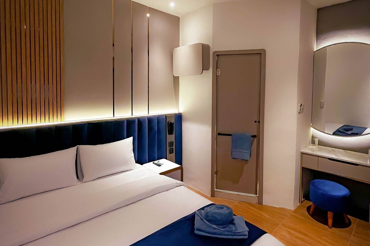 A modern hotel room with a large bed, a bedside table, a bathroom with a mirror, and a wooden floor.