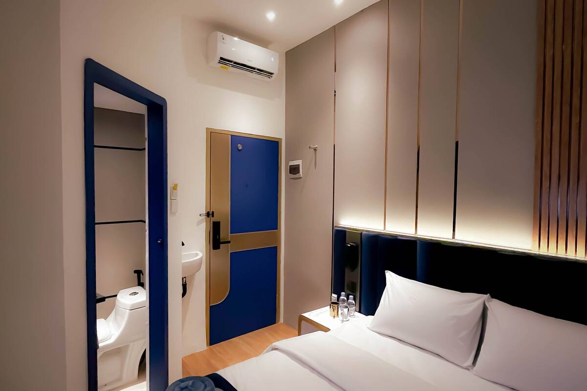A hotel room with a bed, a bedside table with a lamp, a wall-mounted air conditioner, and a door leading to a bathroom.