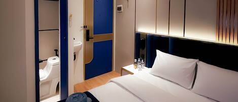 Une chambre d’hôtel comprenant un lit, une table de chevet avec une lampe, un climatiseur fixé au mur et une porte donnant accès à une salle de bain.