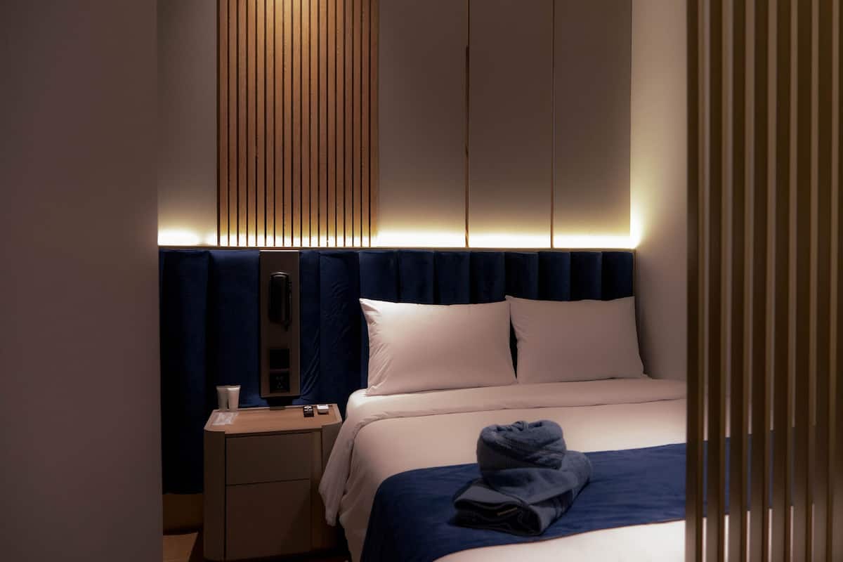 A modern hotel room with a neatly made bed, a bedside table with a lamp, and a large window with vertical blinds.