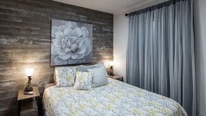 A bedroom with a wooden wall, a large floral painting, a bed with floral bedding, two bedside lamps, and a window with curtains. - Cozy Condo in Ruidoso ideal for Outdoor Enthusiasts!  (Ruidoso)