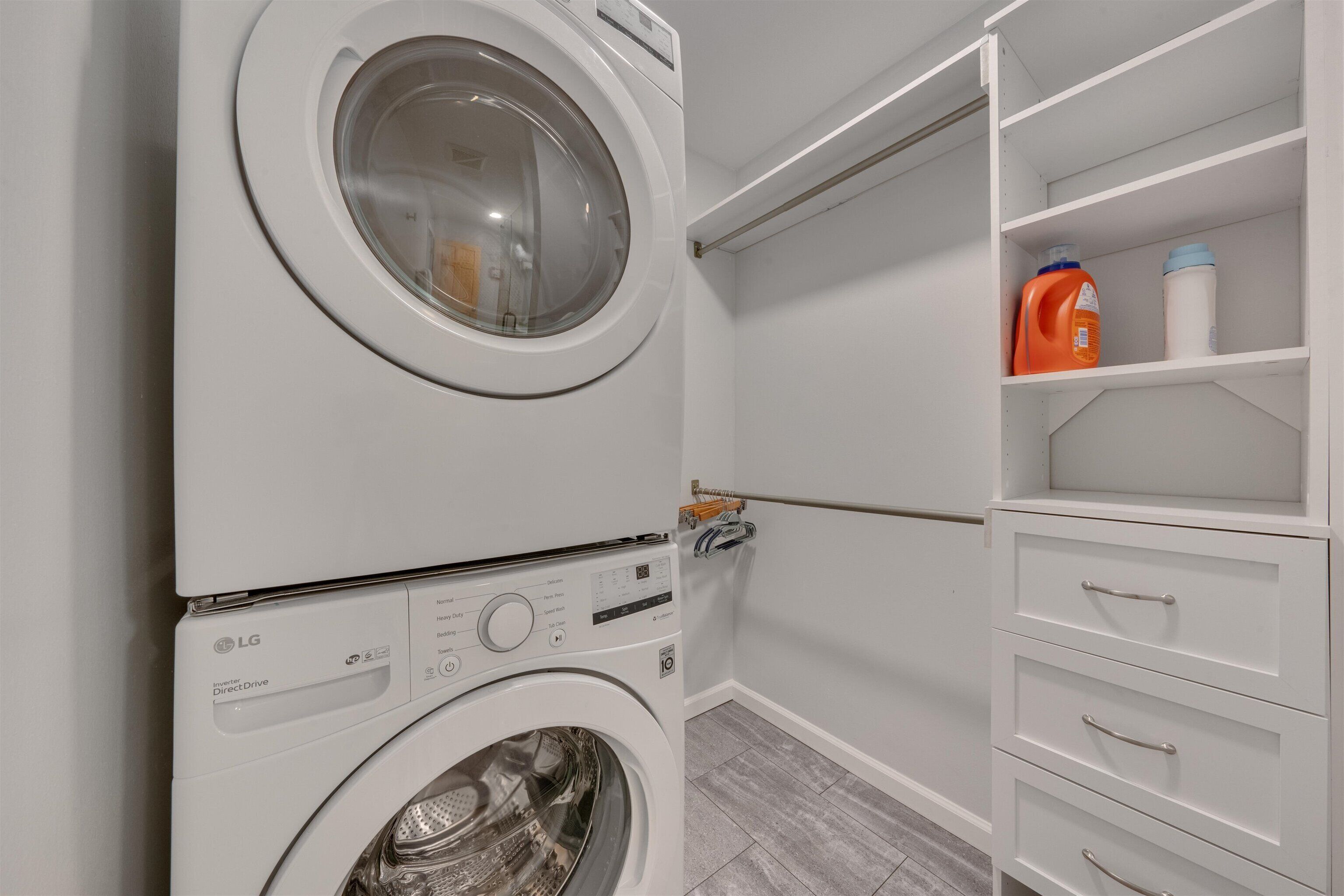 A laundry area with a front-loading washer and dryer, a shelving unit with detergent bottles, and a white storage cabinet.