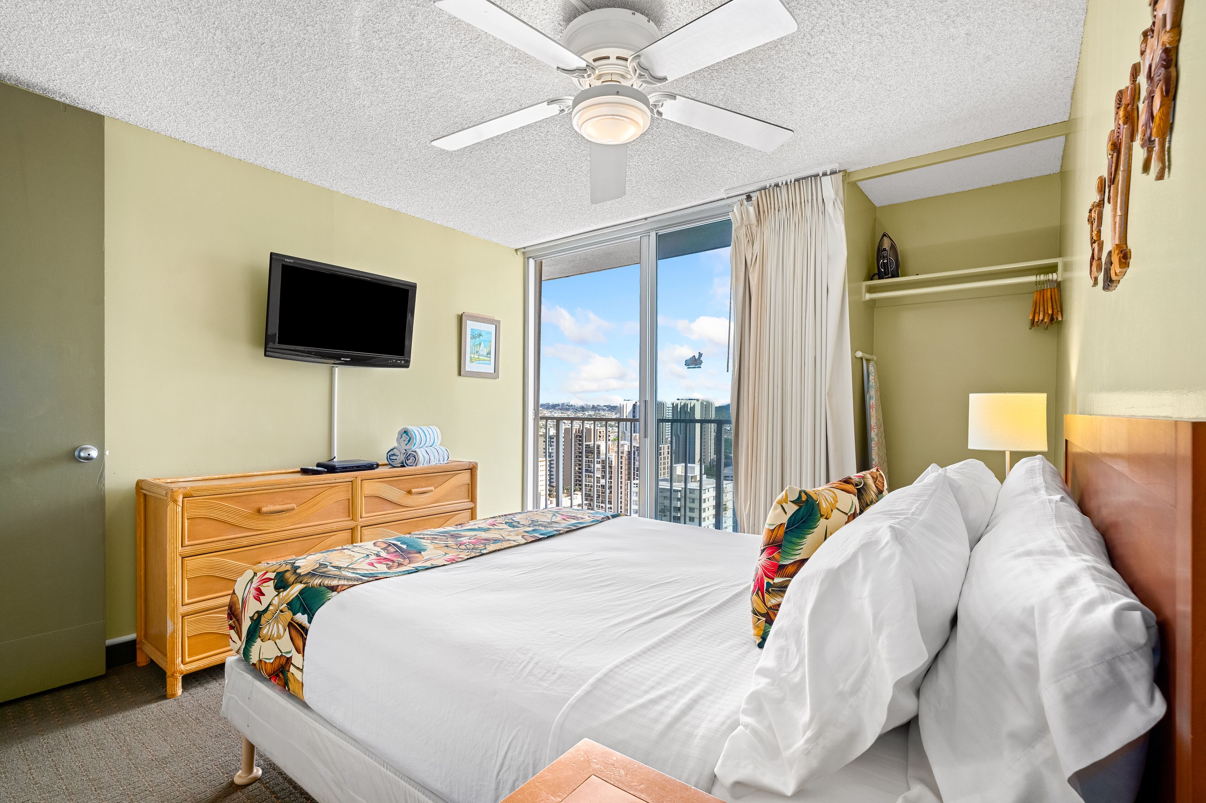A hotel room with a bed, a television, a dresser, and a view of the city through a large window.
